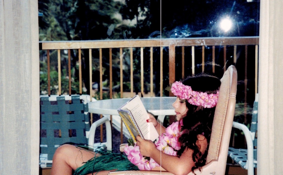 6-year-old Rebecca sits reading in a too-big chair in her hotel room, too engrossed in her book to realize she's being photographed. She wears leis of fake flowers around her head and neck, and a fake grass skirt. Outside the window, palm trees blow in the wind.