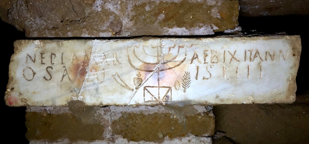 A marble grave-marker set into a brick wall below-ground is inscribed with an epitaph in Latin to either side of a large engraved menorah and other Jewish symbols.