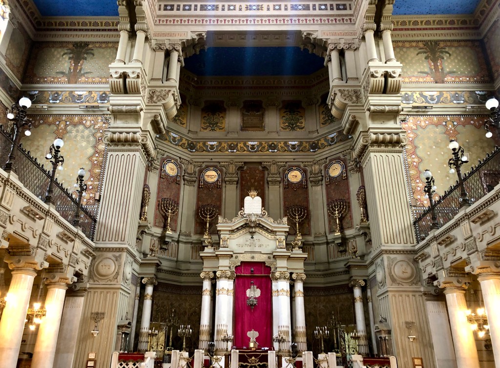 The interior of the Great Synagogue of Rome, lavish with Greek columns, golden menorahs, and frescoes of palm trees and decorative patterns on the walls.