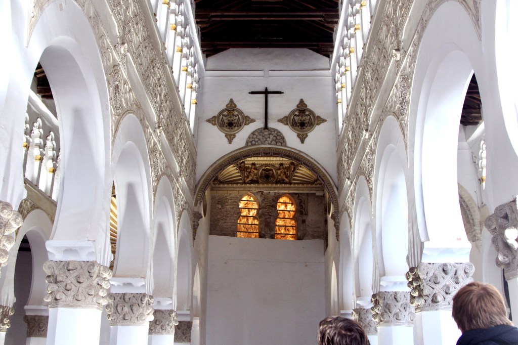 A white-walled synagogue in Moorish style with rows of rounded archways over a colonnade. Above where the ark would have been hangs a cross.