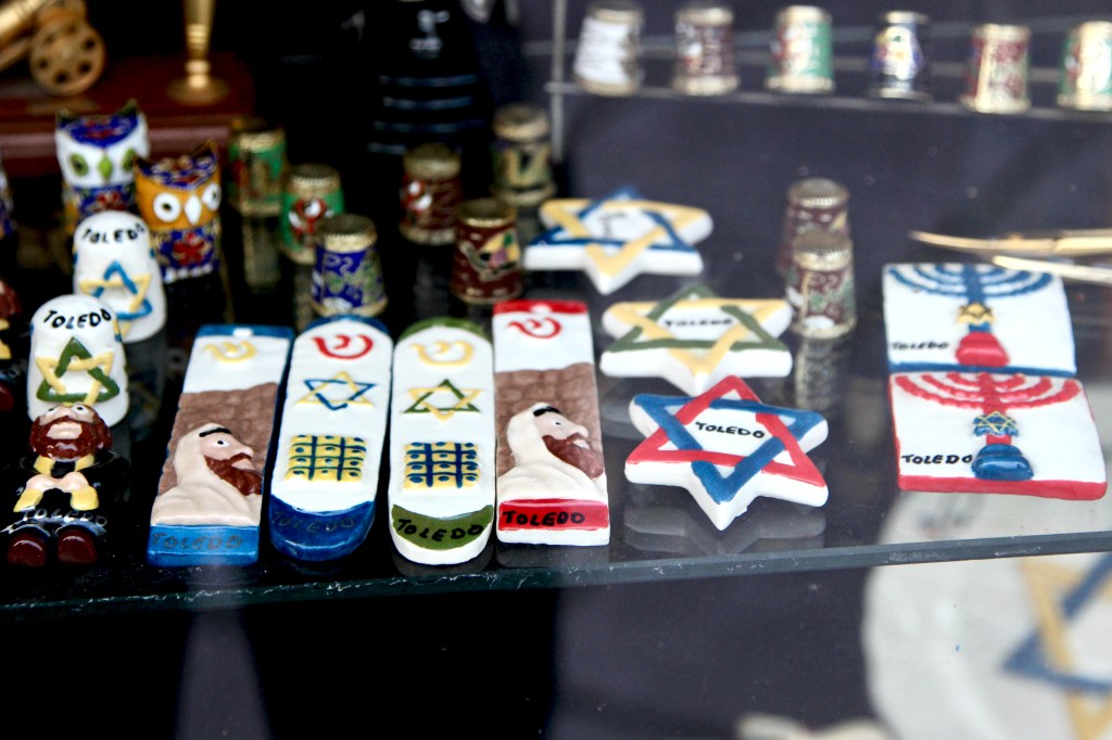 A shop window display showcases ceramic Judaica souvenirs, including klaf-less mezzuzahs. Some of them feature painted rabbis.