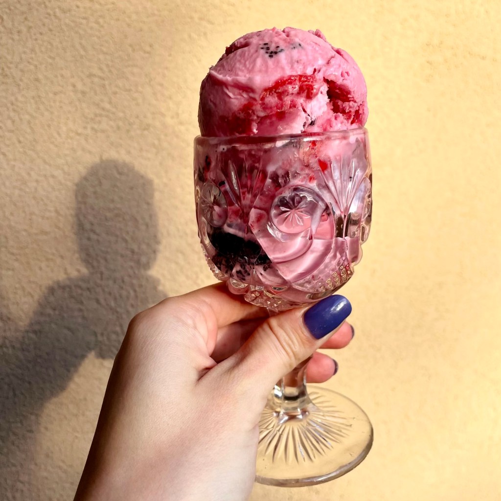 A hand raises a crystal goblet of beet-pink ice cream, with swirls of raspberry jam and poppyseed filling.