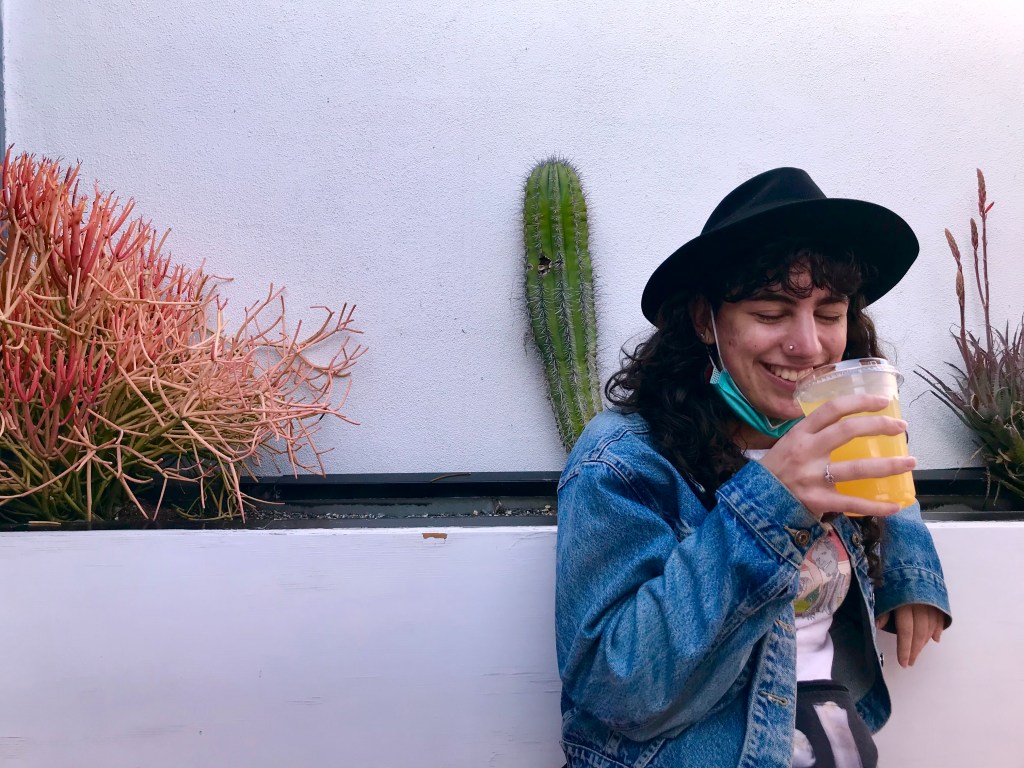 A young woman in a denim jacket smiles as she sips from a cup of ginger-turmeric lemonade. Behind her, set into the wall, is a planter full of cacti (which is how you know this place was built for millennials).