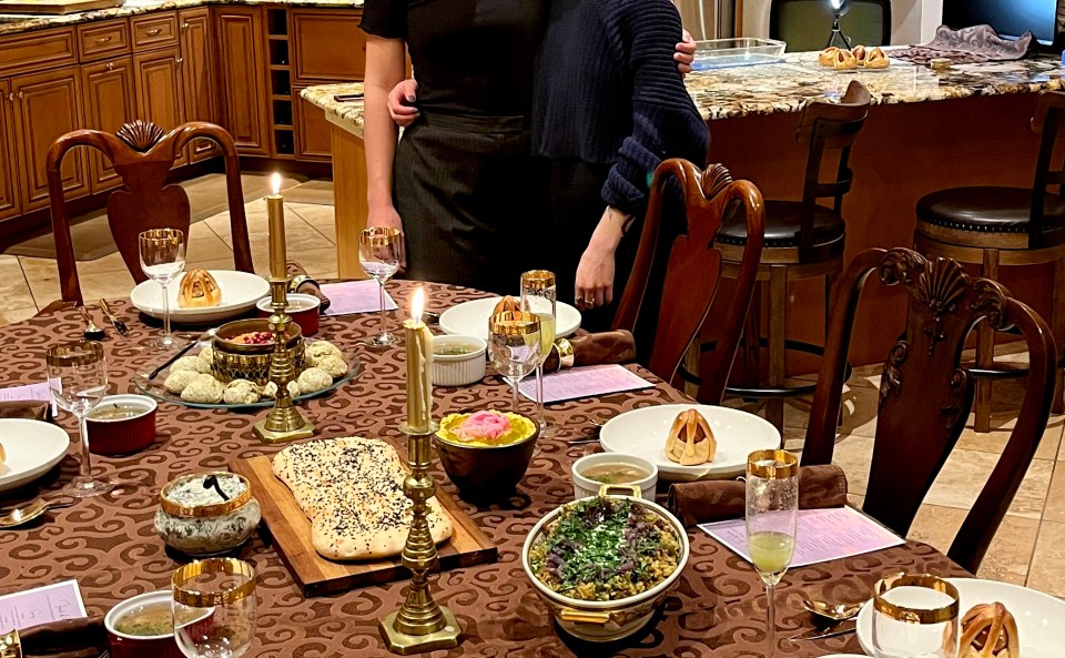 Two tired but smiling cousins stand behind a dining table laden with all the delicious Purim treats they've cooked: foulares, sambusak, naan, mujaddara, hummus, borani, muhammara, eier kichlach, and minestra with orecchiette.