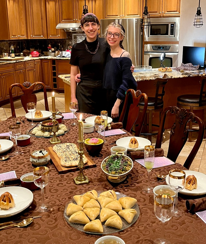 Two tired but smiling cousins stand behind a dining table laden with all the delicious Purim treats they've cooked: foulares, sambusak, naan, mujaddara, hummus, borani, muhammara, eier kichlach, and minestra with orecchiette.