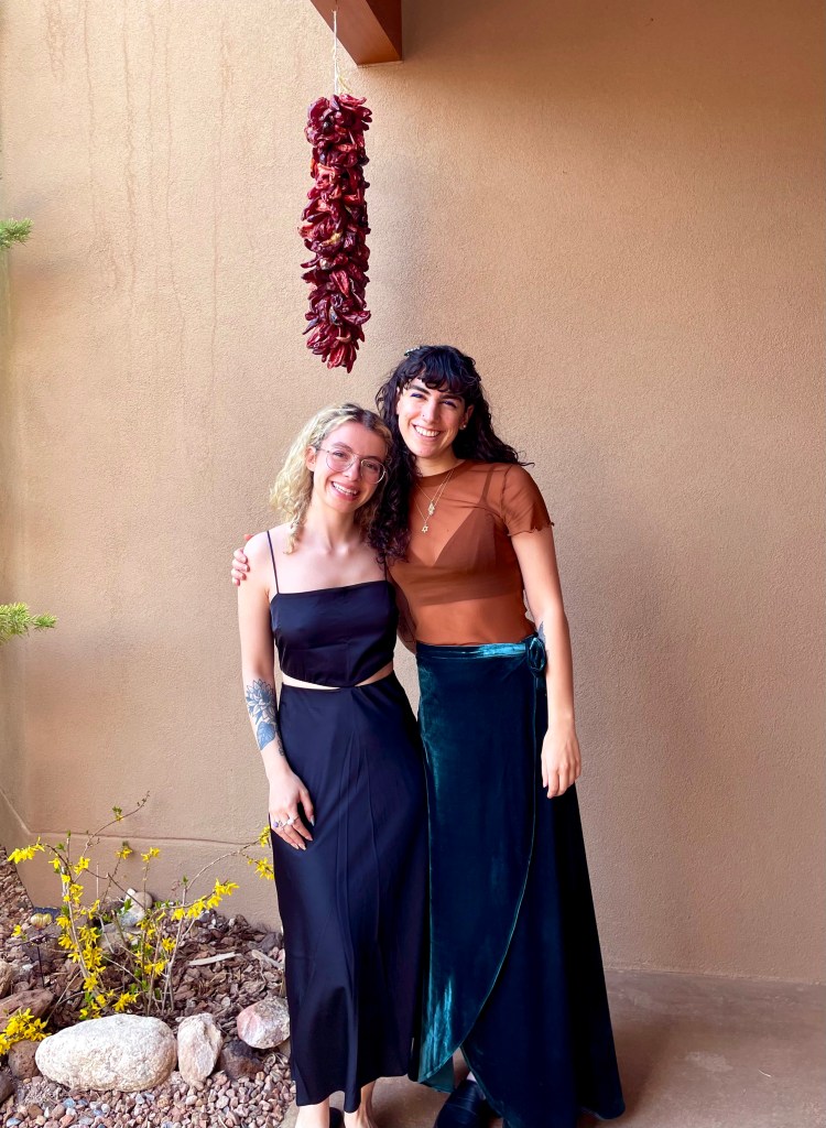 Two young women smile with their arms around one another. They've changed from their kitchen clothes into nice dress for the Seder and are proud of a job well done.