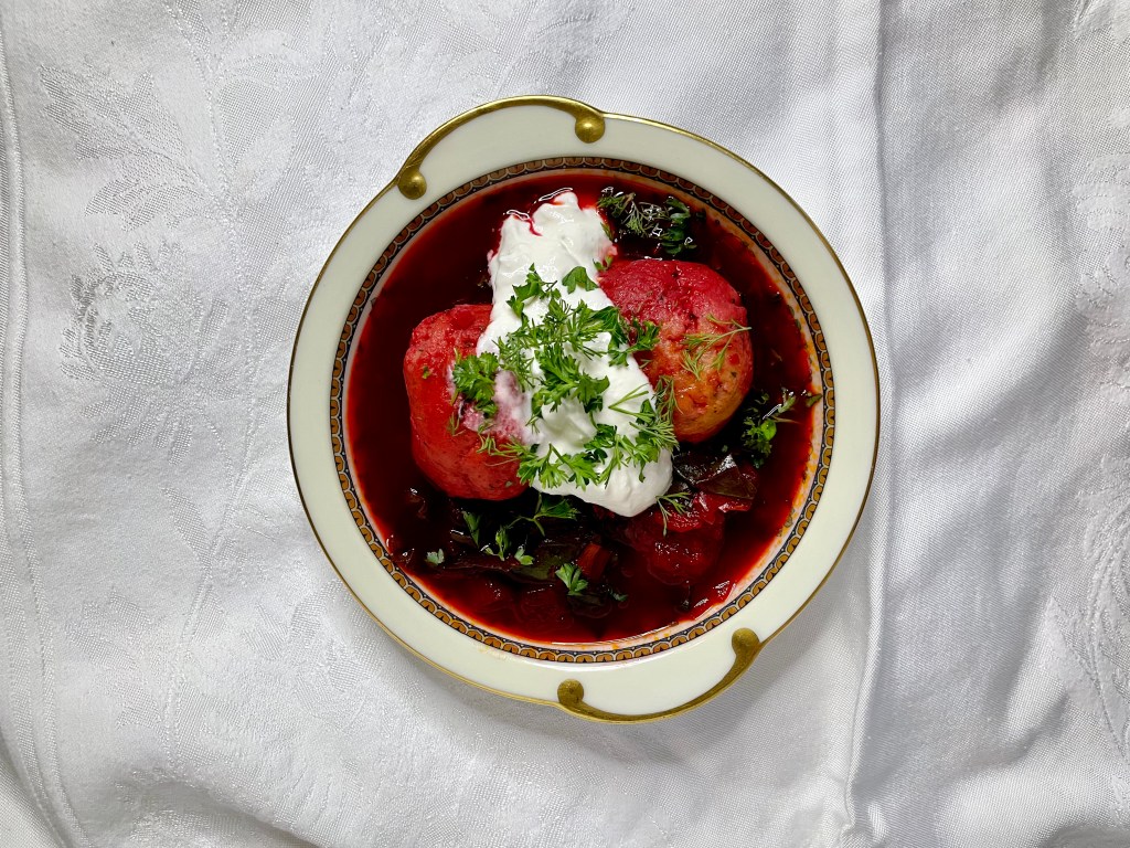 A bowl of matzo ball borscht: beet soup with two fluffy matzo balls and a healthy dollop of horseradish cream and fresh parsley.