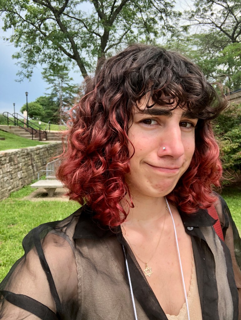 A young woman with pink curly hair takes a very awkward selfie outdoors on her grad school campus. She's showing off how much the humidity has helped her curls but can't take herself seriously enough to smile.