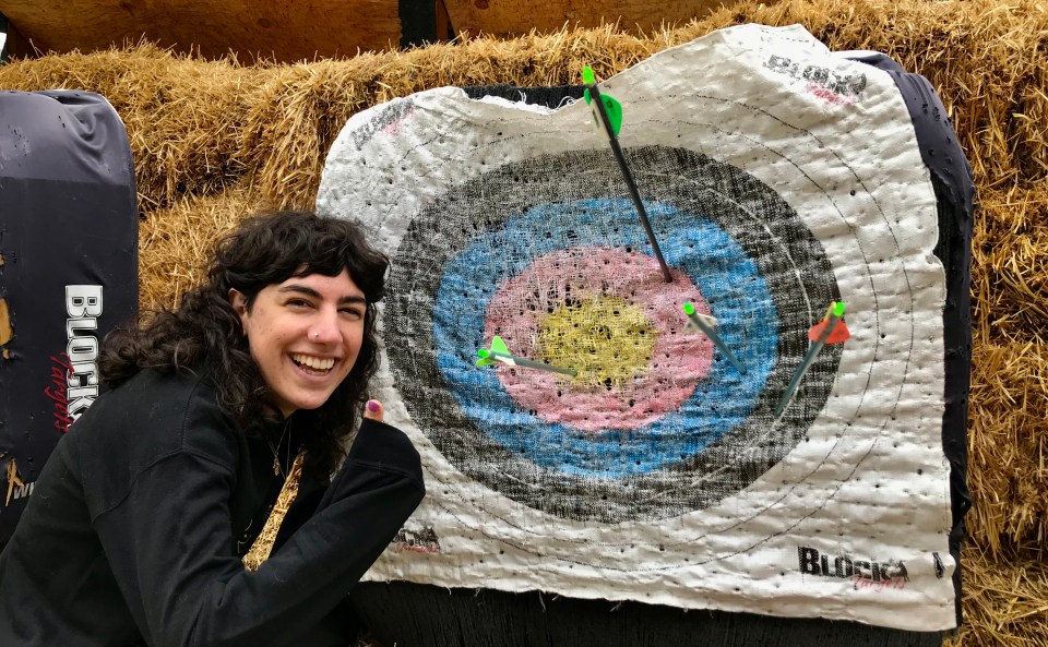 A young woman crouches next to an archery target, giving a thumbs-up. She's hit the target with four arrows, one in each of the four concentric rings. The centermost is a bullseye.