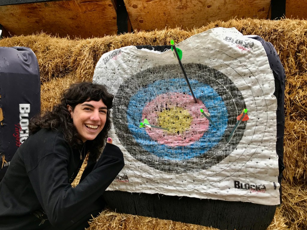 A young woman crouches next to an archery target, giving a thumbs-up. She's hit the target with four arrows, one in each of the four concentric rings. The centermost is a bullseye.