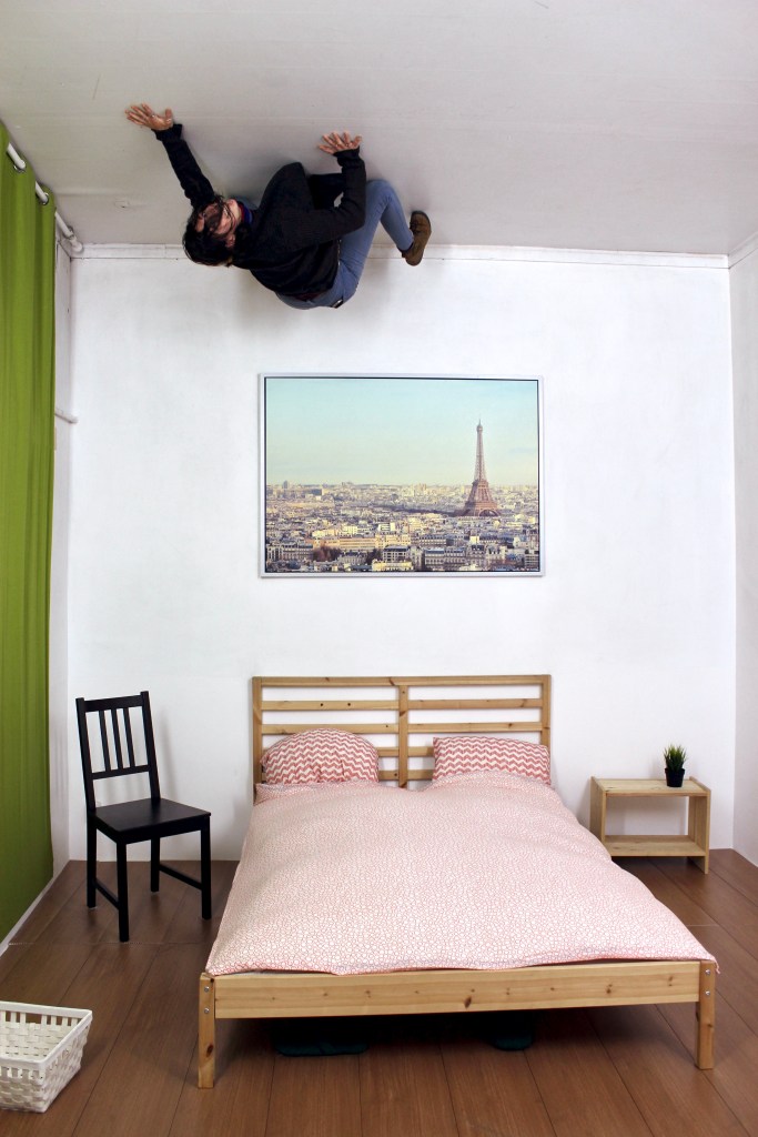 A young woman appears to crawl on the ceiling of a bedroom; the picture is inverted and the furniture is actually attached to the ceiling.