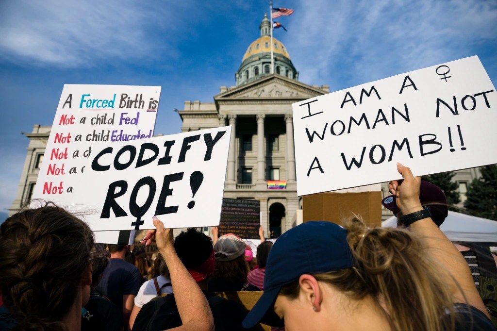 Two people hold up protest signs in a crowd in front of the Colorado State Capitol the weekend after Roe v. Wade was overturned. One reads "Codify Roe!" and the other reads "I am a woman not a womb!!" A third sign is partially visible in the background, with the incomplete text "A Forced Birth is: Not a child Fed, Not a child Educated, Not a ch—"