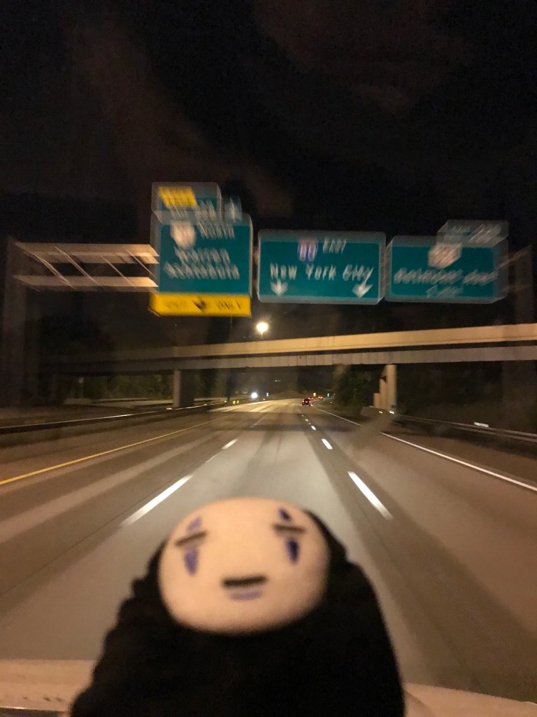 A plushie of the character No Face from the movie Spirited Away lies on the U-Haul dashboard at night as the truck seems to hurtle toward a highway sign pointing toward New York City. Because of the low light at night and the poor camera quality, the signs are blurred in a way that looks like No Face is traveling at warp speed.
