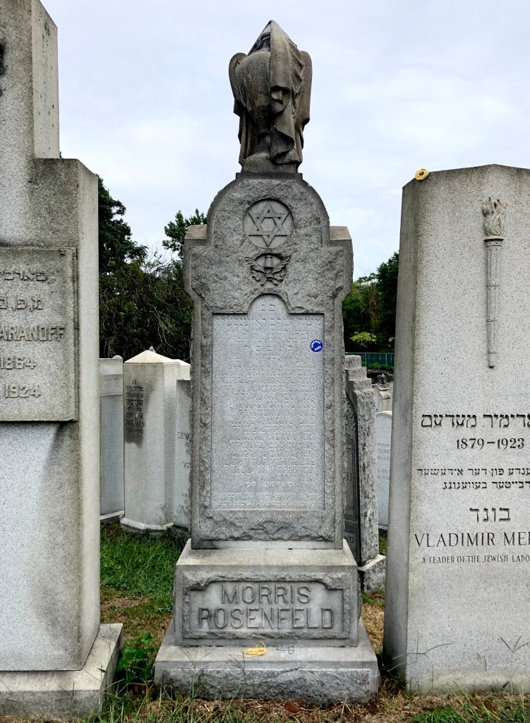 A tall headstone inscribed with the name "Morris Rosenfeld" at the base; the center text contains the epitaph in Yiddish under a Star of David. The whole thing is topped by a carving of an urn draped in a shroud. A small coil of candle wicking rests on the base.